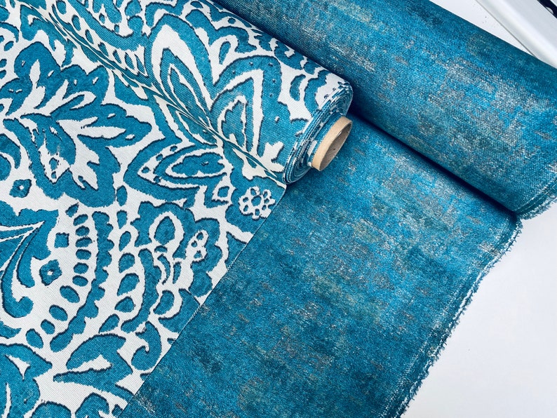 May include: A roll of teal fabric with a white and teal paisley pattern. The fabric is folded in half and the roll is on top of a flat piece of the same fabric.