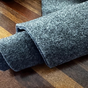 May include: A folded section of grey fabric with a textured surface. The fabric is a medium grey colour and appears to be a heavy material, possibly wool or a similar textile. The fabric is lying on a wooden surface.