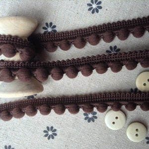May include: Brown pom-pom trim on a wooden spool with three beige buttons. The trim is a decorative fabric with small, round pom-poms.
