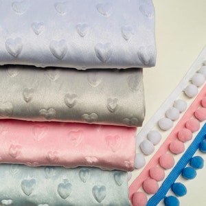 Könnte beinhalten: Vier verschiedene Farben von Stoff mit einem Herzmuster. Der Stoff ist weich und plüschig. Es gibt vier verschiedene Farben von Pompon-Besatz.