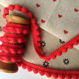 May include: A spool of red pom-pom trim with a wooden centre. The trim is on a beige fabric with a pattern of red hearts and black flowers.