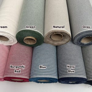 Pode incluir: Uma coleção de rolos de tecido em várias cores, incluindo creme, verde, natural, cinza, rosa blush, vermelho bordô, azul, azul marinho e preto. Cada rolo apresenta um padrão de listras horizontais. Os nomes das cores estão rotulados nos rolos.