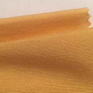 Può includere: Primo piano di un tessuto giallo con una sottile trama tessuta. Il tessuto ha una superficie liscia e leggermente lucida.