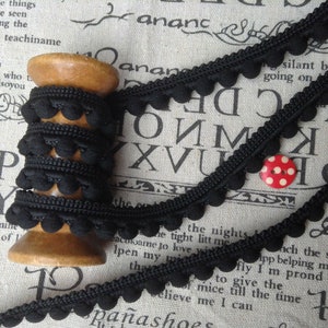 May include: A spool of black pom-pom trim with a wooden bobbin. The trim is made of black fabric with small, round pom-poms. The trim is on a white background with black letters.