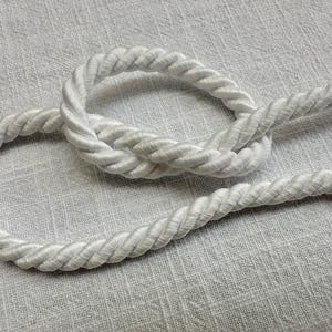 May include: A thick, white twisted rope cord featuring a smooth, satin-like finish. The cord is arranged in a loose, circular loop, showcasing the uniform, spiraled texture of the braided strands throughout the entire length of the material.