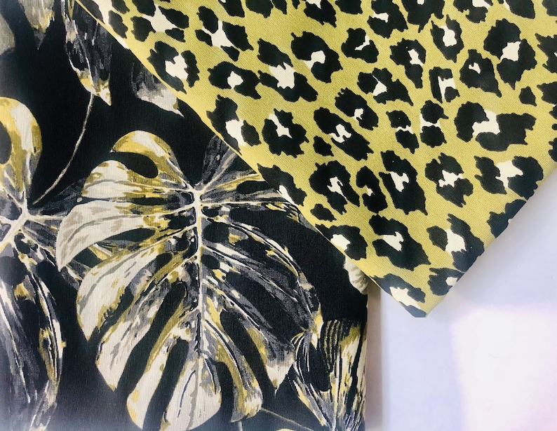 May include: Black fabric with a large white and gold tropical leaf print. The fabric is folded over a green and black leopard print fabric.