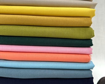 Op de afbeelding: Een stapel van 14 verschillende gekleurde stofstalen. De kleuren zijn wit, beige, geel, mosterd, groen, donkergroen, roze, oranje, lichtblauw, blauw, grijs, bruin en zwart.