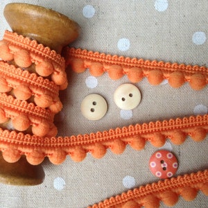 May include: A spool of orange pom-pom trim with a wooden spool. The trim is on a white background with orange polka dots.