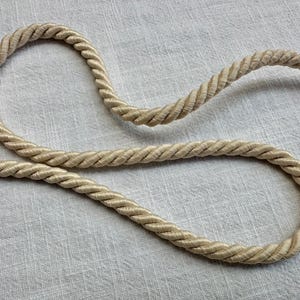 May include: A single length of beige twisted cord featuring a thick, multi-strand rope construction. The cord is arranged in a loose, curved shape, showcasing the uniform texture and consistent light tan color of the twisted fibers throughout.