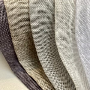 May include: Five different swatches of woven fabric in various shades of gray, beige, and white. The fabric has a loose weave and a slightly textured surface.