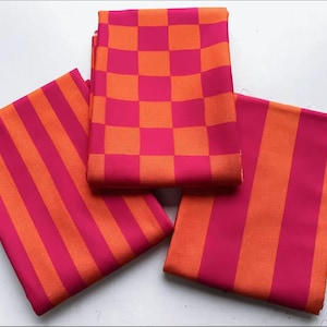 Puede incluir: Tres piezas de tela dobladas en colores vibrantes. Dos piezas presentan un patrón audaz de rayas naranjas y fucsias alternas. La tercera pieza muestra un diseño de tablero de ajedrez de los mismos colores. Las telas están cuidadosamente dispuestas, mostrando los patrones contrastantes.