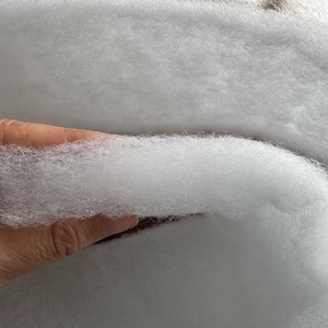 May include: Close-up of fluffy, white polyester fiberfill. The soft, cloud-like material is held in a hand, showcasing its texture. This filling is commonly used for pillows, cushions, and crafts.