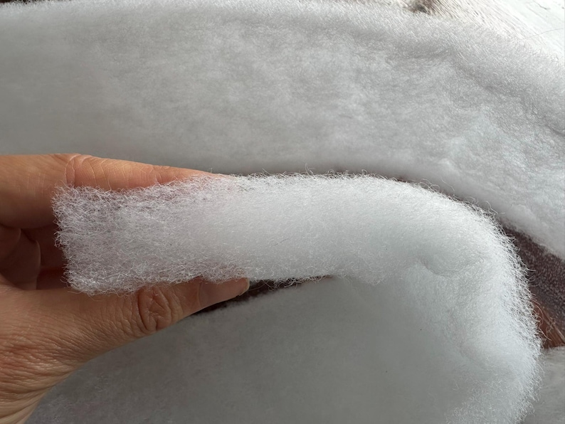 May include: Close-up of fluffy, white polyester fiberfill. The soft, cloud-like material is held in a hand, showcasing its texture. This filling is commonly used for pillows, cushions, and crafts.