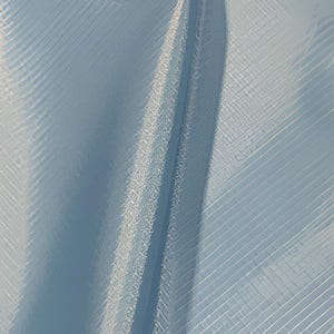 Può includere: Primo piano di un tessuto azzurro con un motivo a trama diagonale strutturato. Il tessuto ha una lucentezza sottile e una cucitura verticale al centro, che crea ombre e riflessi.