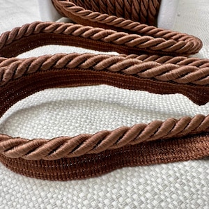 May include: Close-up of brown braided trim with a textured surface. The trim is folded in half, showing the underside of the braid.