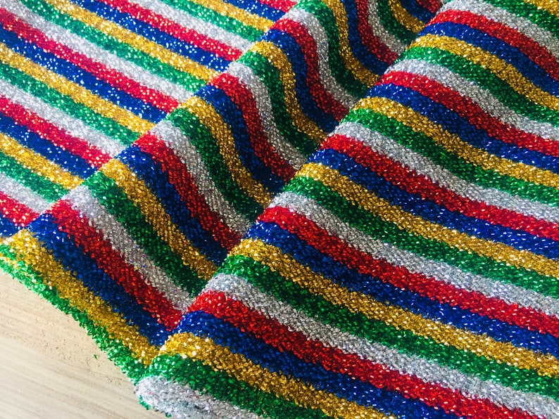 May include: A close-up of a fabric with a striped pattern in red, blue, green, gold, and silver. The fabric is shiny and has a glittery texture.