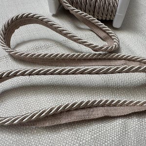 May include: A close-up of a beige braided trim with a soft, textured surface. The trim is folded in half, showing the inside and outside of the braid.