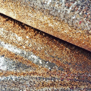 Op de afbeelding: Close-up van een stof bedekt met kleine, glanzende, gouden pailletten. De pailletten zijn dicht op elkaar gepakt, waardoor een glinsterend effect ontstaat. De stof is gevouwen en onthult een donkere, contrasterende onderkant. De algehele textuur is rijk en luxueus.