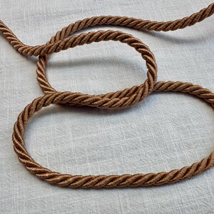 May include: A length of medium brown twisted cord featuring a multi-strand braided construction. The cord is arranged in a loose, overlapping loop pattern, showcasing the uniform texture and consistent thickness of the twisted fibers throughout.
