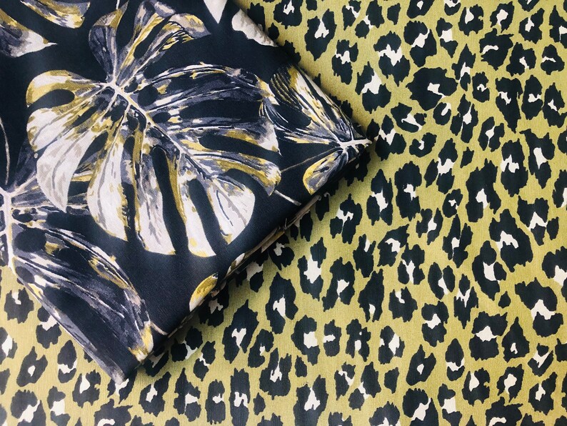 May include: Two fabric swatches with different patterns. One swatch features a black background with large, gold and white tropical leaves. The other swatch features a green background with a black leopard print.