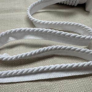 May include: White rope trim with a twisted rope design. The trim is made of a soft, flexible material and is ideal for adding a decorative touch to clothing, home decor, or craft projects. The trim is shown on a neutral background.