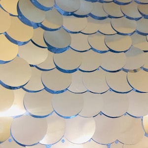 Könnte beinhalten: Nahaufnahme einer schimmernden Oberfläche, die aus sich überlappenden, kreisförmigen, metallisch goldenen Pailletten besteht. Die Pailletten sind in einem wellenartigen Muster auf einem hellblauen Hintergrund angeordnet und erzeugen einen strukturierten, reflektierenden Effekt. Die Gesamtästhetik ist glamourös und dekorativ.
