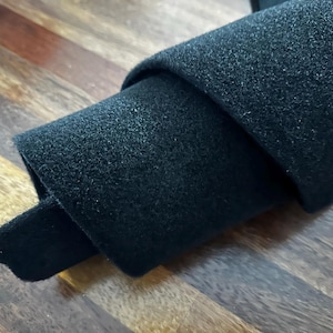 May include: A rolled-up piece of black felt fabric. The felt has a soft, textured surface and is partially unrolled, revealing the inner layers. The fabric is lying on a wooden surface.