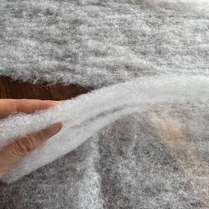 May include: Close-up of a hand holding a section of white, fluffy batting material. The batting is layered over a larger piece of gray, textured fabric. The material appears soft and lightweight, suitable for various craft projects.
