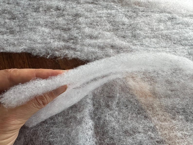 May include: Close-up of a hand holding a section of white, fluffy batting material. The batting is layered over a larger piece of gray, textured fabric. The material appears soft and lightweight, suitable for various craft projects.