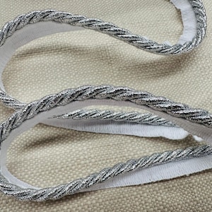 May include: Silver cord trim with a white backing. The cord is twisted and has a metallic sheen.