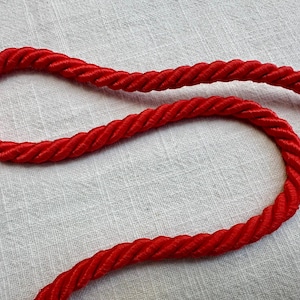 May include: A vibrant red twisted cord made of multiple strands of synthetic fiber. The rope features a smooth, glossy finish and a consistent, tightly coiled structure that curves across the frame in a gentle, flowing S-shaped pattern.