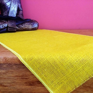 May include: A yellow hessian table runner with a white border. The runner is on a wooden surface and a small, dark grey statue of a Buddha is sitting in front of it.
