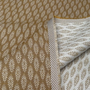 May include: A folded fabric with a repeating leaf pattern. The fabric is a warm, golden-brown colour with a white leaf design. The edge of the fabric has a striped border.