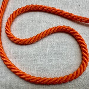 May include: A bright orange twisted cord featuring a thick, multi-strand rope construction with a smooth, glossy finish. The cord is arranged in a curved, looping pattern, showcasing the uniform spiral texture of the twisted fibers throughout.
