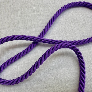 May include: A single length of vibrant purple twisted cord arranged in a loose, overlapping loop. The cord features a distinct, tightly wound rope construction with a smooth, slightly reflective sheen along its entire length.