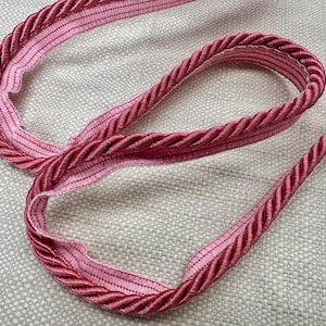 May include: A length of decorative trim in shades of pink and red. The trim features a twisted rope design on the outer edge and a lighter pink fabric on the inner edge. The trim is lying on a textured, light-colored surface.