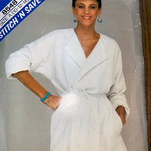 Könnte beinhalten: Ein weißer Bademantel mit Schalkragen und Bindegürtel. Der Bademantel hat an jeder Seite eine Tasche. Die Modellnummer 8548 und der Text "Stitch 'N Save" sind auf der Verpackung sichtbar.