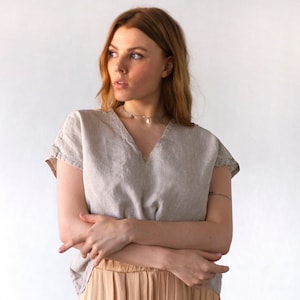 以下が含まれることがあります： Vネックと半袖のナチュラルグレーのリネン素材のトップスを着た女性。薄い茶色のスカートと小さなチャームのついたシルバーネックレスをしています。