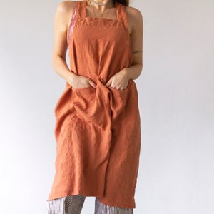 Peut inclure: Un tablier en lin terracotta avec des bretelles réglables et deux grandes poches. Le tablier est porté par une personne, mais l'accent est mis sur le tablier lui-même.