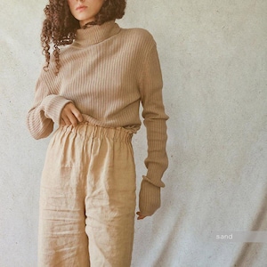 May include: A beige ribbed knit turtleneck sweater and matching linen pants. The long-sleeved sweater has a high neck and fitted cuffs. The pants have a gathered waistband. The word "sand" is in the bottom right corner.