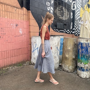 K&ouml;nnte beinhalten: Eine Frau tr&auml;gt ein rostfarbenes Tanktop und einen hellgrauen Leinenrock und geht auf einem gepflasterten Weg. Der Hintergrund zeigt eine Backsteinmauer mit Graffiti und einen Baumstumpf.