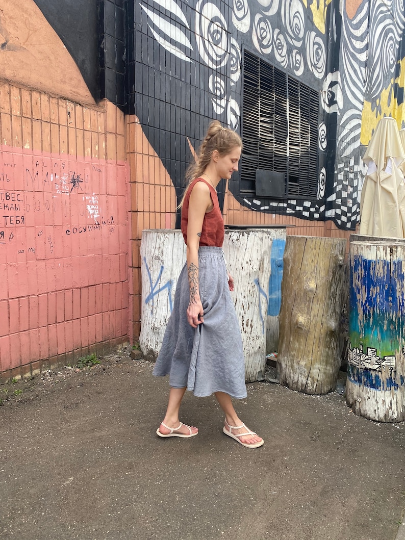K&ouml;nnte beinhalten: Eine Frau tr&auml;gt ein rostfarbenes Tanktop und einen hellgrauen Leinenrock und geht auf einem gepflasterten Weg. Der Hintergrund zeigt eine Backsteinmauer mit Graffiti und einen Baumstumpf.