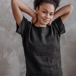 Peut inclure: Une femme aux cheveux boucl&eacute;s fonc&eacute;s porte une chemise en lin noire &agrave; manches courtes. Elle sourit et a les bras lev&eacute;s derri&egrave;re la t&ecirc;te.