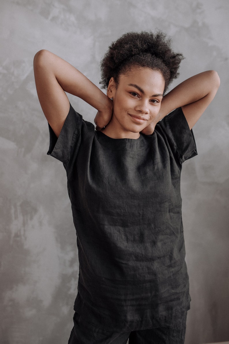 Peut inclure: Une femme aux cheveux boucl&eacute;s fonc&eacute;s porte une chemise en lin noire &agrave; manches courtes. Elle sourit et a les bras lev&eacute;s derri&egrave;re la t&ecirc;te.