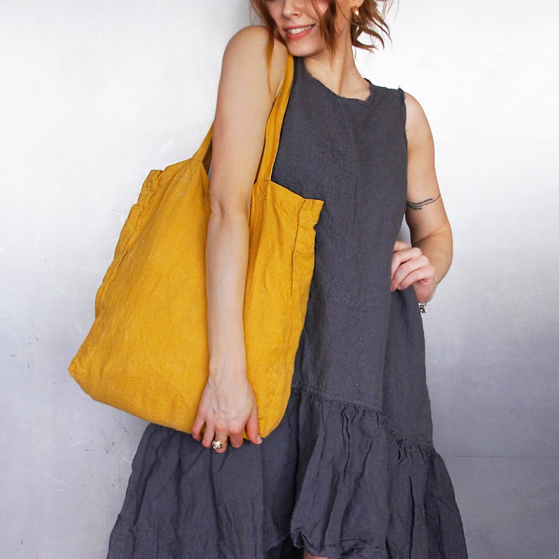 May include: A mustard yellow tote bag with a textured appearance is held by a person wearing a gray sleeveless dress. The dress has a ruffled hemline. The background is a light gray.