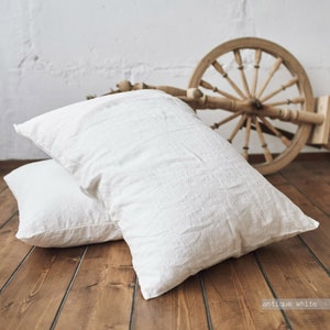 May include: Two white linen pillowcases on a wooden floor. The pillowcases are made of a soft, natural fabric and have a slightly textured surface. The color of the pillowcases is described as "antique white".