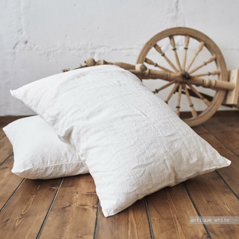 May include: Two white linen pillowcases on a wooden floor. The pillowcases are made of a soft, natural fabric and have a slightly textured surface. The color of the pillowcases is described as "antique white".