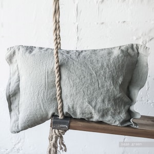 Op de afbeelding: Een saliegroen linnen kussen met een touw dat eraan hangt. Het kussen ligt op een houten schommel.