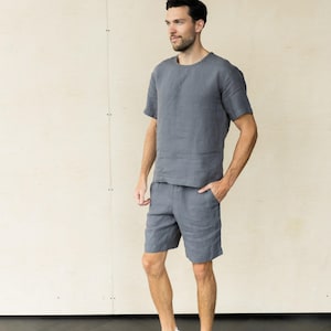 以下が含まれることがあります： グレーのリネン素材の半袖シャツとショートパンツのセット。モデルは白いスニーカーを履いています。