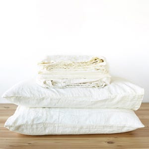 May include: A stack of two rectangular white pillows with a textured fabric finish, topped with a folded white fabric sheet. The items are arranged in a neat pile, showcasing the natural weave and soft appearance of the white textile material.